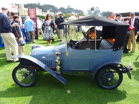 Side view of a pale blue two-seater with a black hood, brass horn and lamps on grass, people standing behind. Opens a larger view