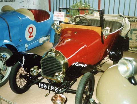 Red two-seater with black wings and brass lamps between a blue racing car and a cream car. Opens a larger view