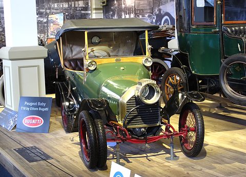 Green two-seater with a black hood, brass radiator and red-spoked wheels beside a Bugatti display sign. Opens a larger view
