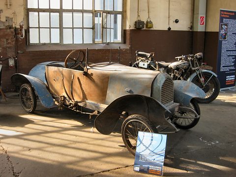 Dusty, faded grey-blue little two-seater with crumpled wings in an old hall, a motorcycle and an information board beside it. Opens a larger view