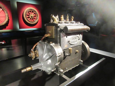 Small four-cylinder engine with brass fittings and a Lion Peugeot plate, on a dark plinth before red panels. Opens a larger view