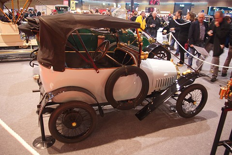 Cream two-seater with a brown folding hood, spare wheel on its side and black wings, visitors behind a rope. Opens a larger view