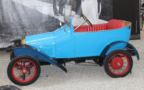 Side view of a bright blue two-seater with red upholstery, red-spoked wheels and black wings on gravel. Opens a larger view
