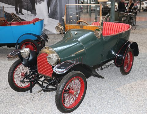 Dark green two-seater with a red radiator, red seats and red wire wheels on grey gravel. Opens a larger view