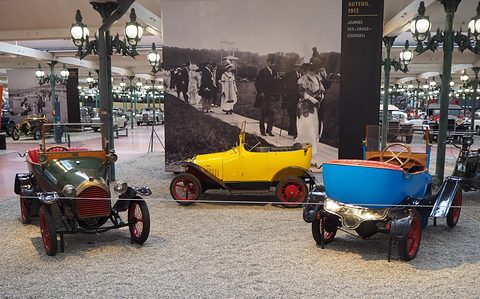 Three small open cars in a row on gravel under lamp posts, a large old photograph on a screen behind. Opens a larger view