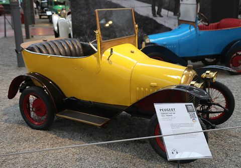 Yellow open two-seater with red-spoked wheels and a museum label standing in front of its wheel. Opens a larger view