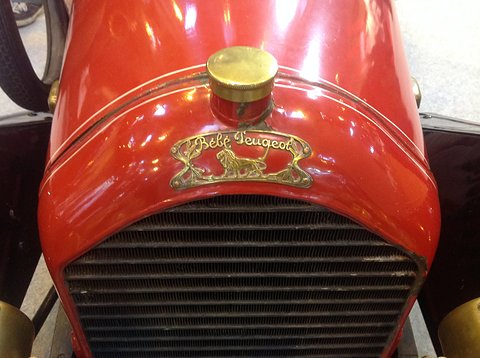 Close-up of a red bonnet with a brass filler cap, a gilt lion badge in script and a radiator grille. Opens a larger view
