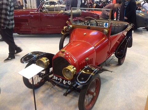 Red two-seater with brass headlamps and black wings, a small information stand in front. Opens a larger view