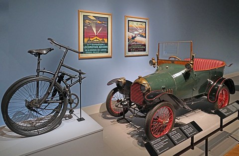 Green two-seater with red wheels on a low plinth, an old military bicycle and two framed posters beside it. Opens a larger view