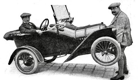 A man in a tweed suit lifts the back of a small open car with a driver aboard. Opens a larger view
