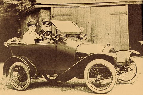A sepia photograph of a small open two-seater with wire wheels and a rounded bonnet, a woman and a man in a cap aboard. Opens a larger view
