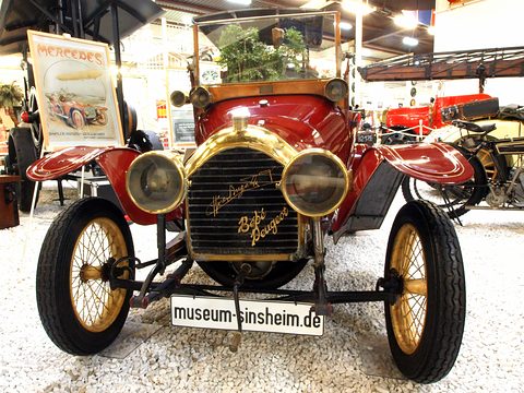 Front of a red two-seater with a brass radiator bearing a Bugatti script, twin brass lamps and a museum plate below. Opens a larger view