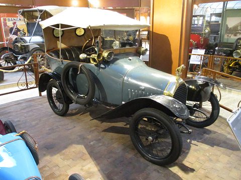 Blue-grey two-seater with a white folding hood, spare wheel and black wings on a brick floor. Opens a larger view