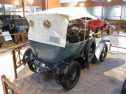 Back of a blue-grey two-seater with a white hood and a rear number plate, behind a decorative railing. Opens a larger view