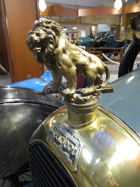 Brass lion figure standing on a radiator cap above an oval Peugeot badge, a blue car behind. Opens a larger view
