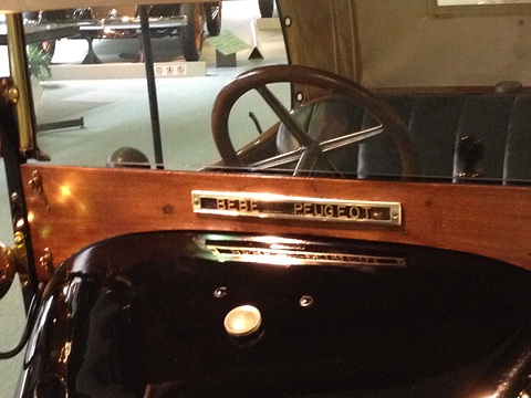 Varnished wooden dash with a brass nameplate, a black scuttle and a wooden steering wheel behind. Opens a larger view
