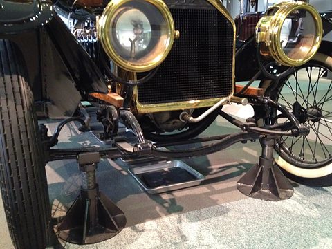 Low close view of a beam axle, springs, brass lamps and a grille, the car raised on stands. Opens a larger view