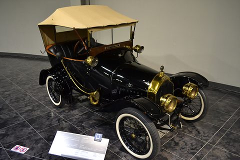 Black two-seater with a cream hood, brass lamps and white-wall tyres on a polished tiled floor. Opens a larger view