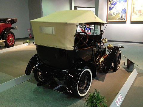 Back of a black two-seater with a cream folding hood and white-wall tyres on a raised display. Opens a larger view