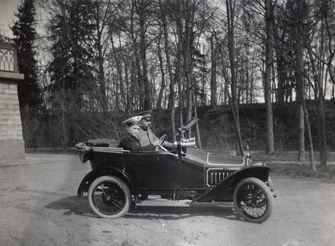 A boy in a cap sits at the wheel of a small dark open car in a winter wood. Opens a larger view