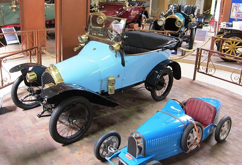 Light blue open two-seater with brass radiator and black wings, a small bright blue child’s racing car in front. Opens a larger view