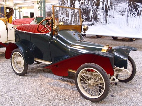 Side view of a dark green two-seater with red wings and red seats before a large old photograph. Opens a larger view