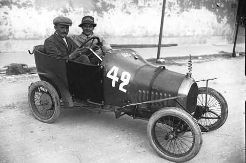 Two men in caps in a narrow dark racing car numbered 42, a coiled spring on its bonnet, on a sunny road. Opens a larger view