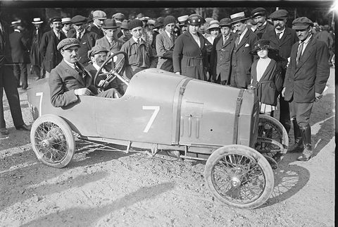 A man at the wheel of a pale pointed-tail racing car numbered 7, a crowd of men in hats and caps behind. Opens a larger view