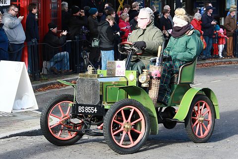 Green veteran car with red wheels carrying two people in warm coats, spectators lining the street. Opens a larger view