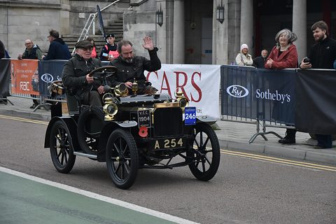 Black veteran two-seater with brass lamps, two men in caps aboard, crowd and banners behind. Opens a larger view