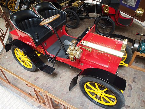 High view of a red veteran car with brass bonnet trim, two black seats and yellow wheels. Opens a larger view