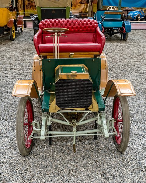 Front of a small green veteran car with brass trim, a red buttoned seat and red wheels on pebbles. Opens a larger view