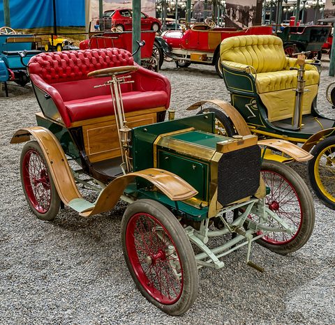 Green and varnished-wood veteran car with red buttoned seats and red wheels on grey pebbles. Opens a larger view
