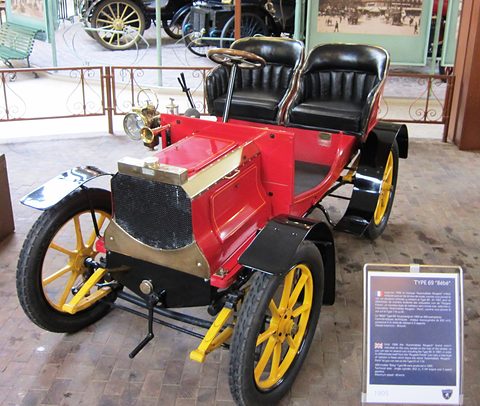 Red veteran two-seater with yellow wooden wheels, two black buttoned seats and a museum panel beside it.