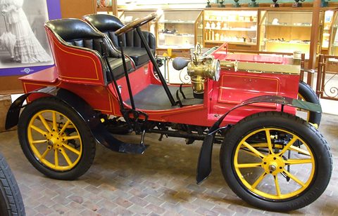 Side view of a red veteran two-seater with yellow wooden wheels, brass lamp and black seats. Opens a larger view