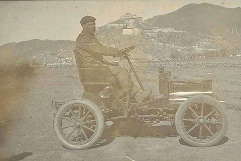 A faded photograph of a man in a cap at the wheel of a tiny veteran car with wooden wheels, a hilltop fortress behind. Opens a larger view