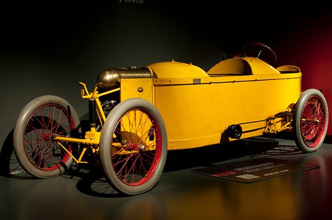 Side view of a long low yellow cyclecar with red wire wheels in dark museum lighting. Opens a larger view
