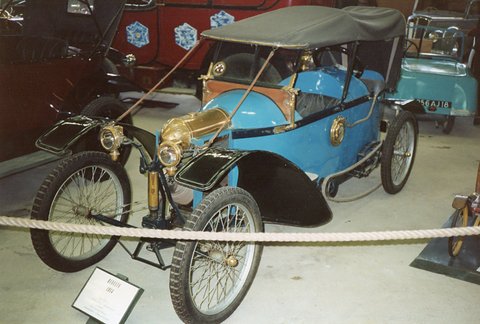 Blue narrow cyclecar with a black hood and brass lamps behind a rope barrier. Opens a larger view