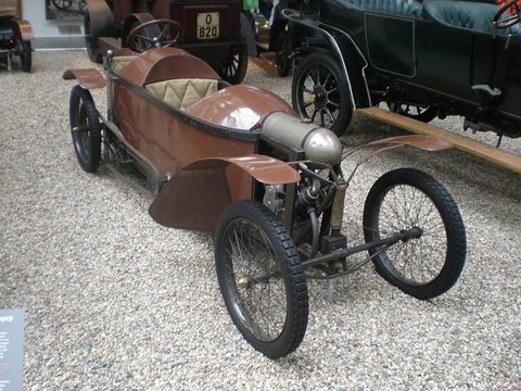 Narrow brown wooden-bodied cyclecar with cycle wings and an exposed engine on gravel among veteran cars. Opens a larger view