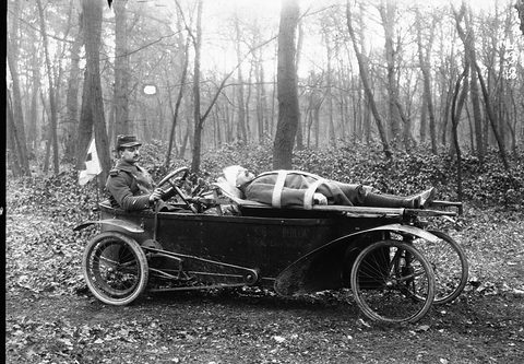 A soldier sits at the back of a narrow open car while a wounded man lies on a stretcher along its body.