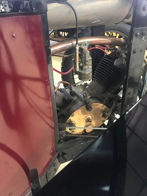 Close view of a V-twin engine with finned cylinders inside a maroon cyclecar body beside a wire wheel. Opens a larger view