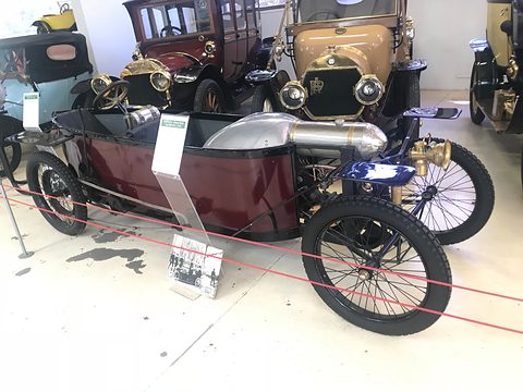 Maroon narrow cyclecar with a long bonnet and wire wheels behind a red rope, a label in front. Opens a larger view