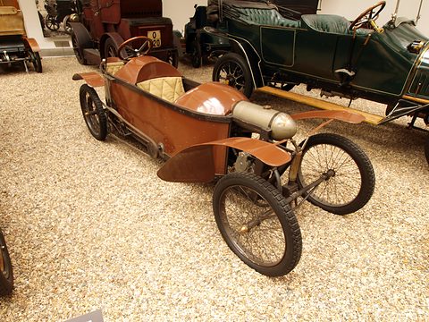 Boat-like brown wooden cyclecar with tandem seats, cycle wings and wire wheels on gravel. Opens a larger view
