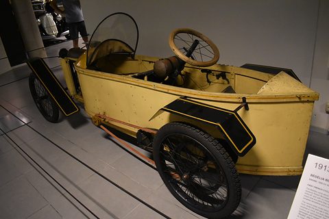 Rear of a yellow narrow cyclecar with a steering wheel at the back seat and a pointed tail. Opens a larger view