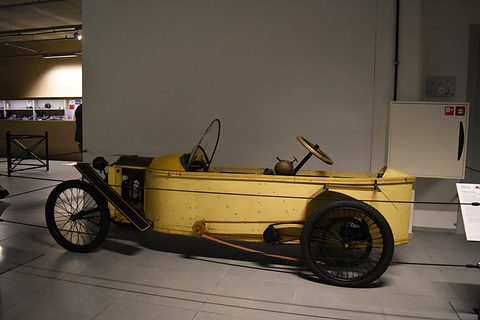 Side view of a long narrow yellow cyclecar with wire wheels on a grey museum floor. Opens a larger view