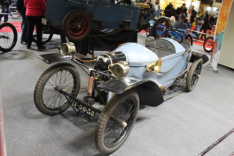Pale blue narrow cyclecar with large brass lamps and wire wheels on grey carpet, visitors behind. Opens a larger view