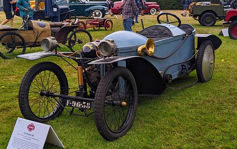 Pale blue narrow cyclecar with brass lamps and wire wheels on grass, a white display board in front. Opens a larger view