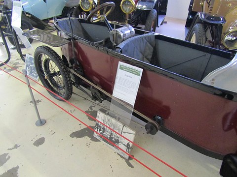 Looking down into a narrow maroon cyclecar with two seats one behind the other and a steering wheel at the back. Opens a larger view