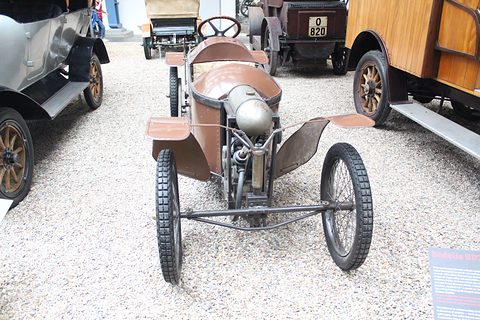 Front of a narrow cyclecar with an exposed V-twin engine between wide cycle wings on gravel. Opens a larger view