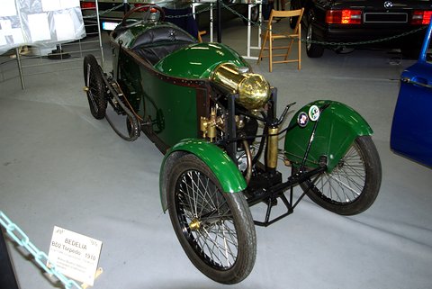 Green narrow cyclecar with brass lamps, cycle wings and wire wheels, a label board beside it. Opens a larger view